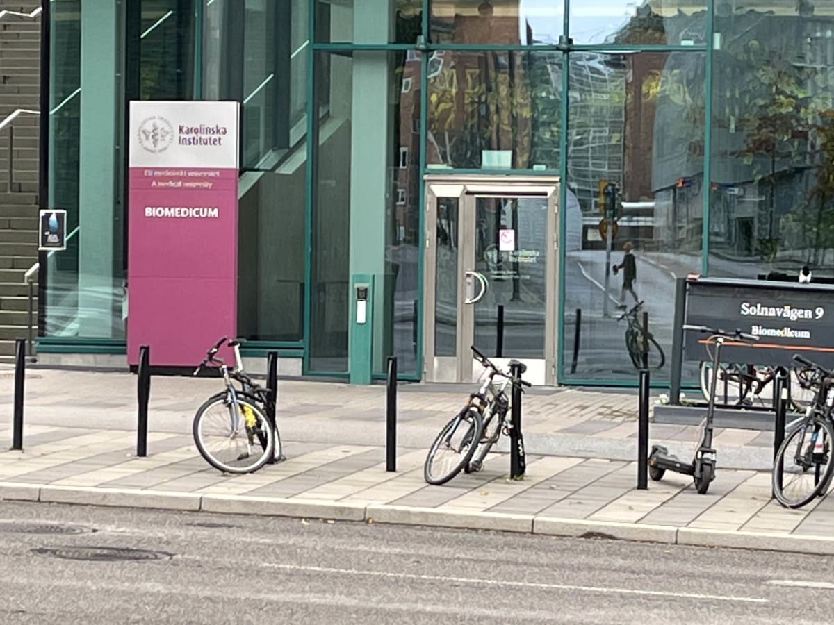 Bilden visar utsidan av en modern byggnad med glasfasad och tegeldetaljer. En stor rosa skylt med texten "Karolinska Institutet" och "BIOMEDICUM" är placerad framför byggnaden. Framför byggnaden finns ett gångstråk med ljusa stenplattor och en asfalterad väg. Flera cyklar är parkerade vid svarta stolpar och ett cykelställ. Till höger i bilden ses en svart skylt med texten "Solnavägen 9" och "Biomedicum". En person är synlig reflekterad i glasfasaden och också svagt synlig gående på avstånd. En elsparkcykel är också parkerad intill cykelstället.