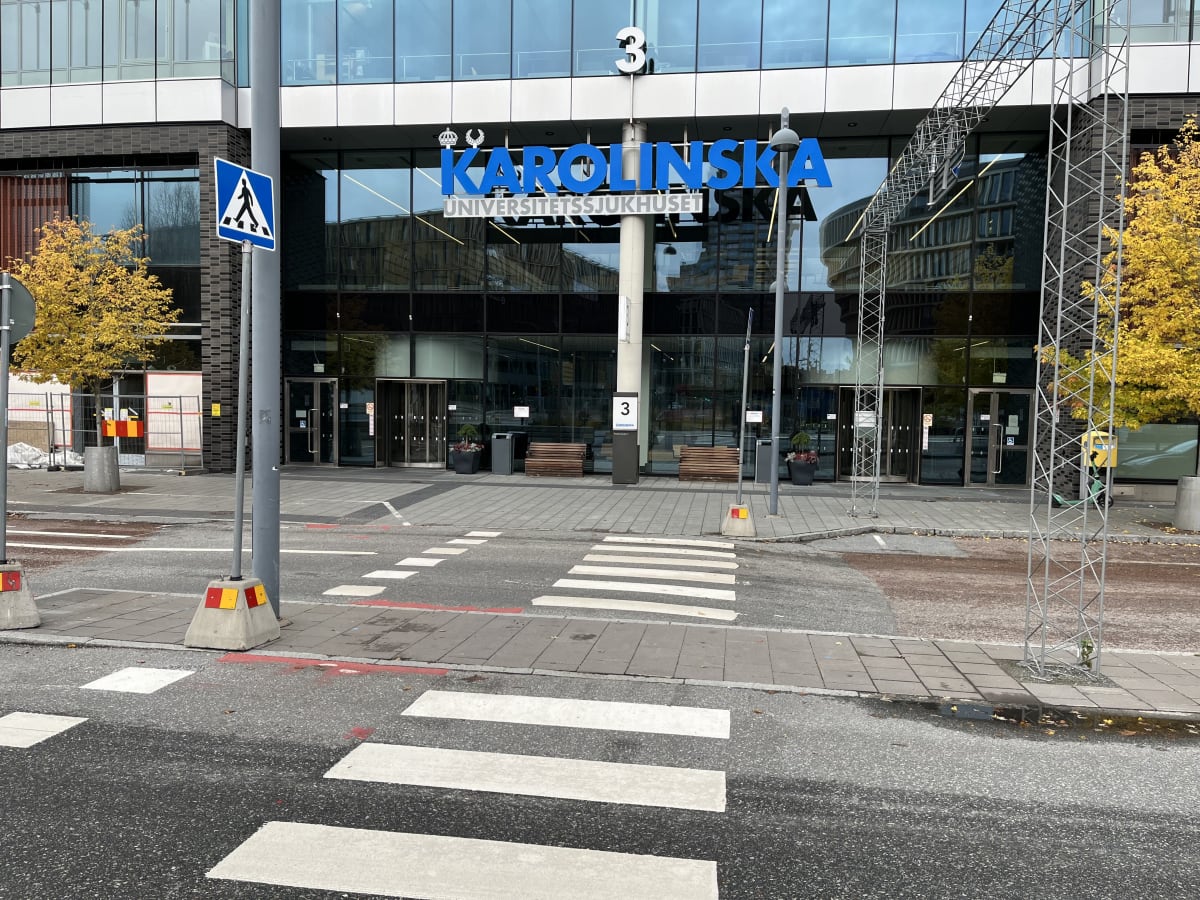 Bilden visar entrén till en modern byggnad, identifierad som Karolinska Universitetssjukhuset, med stora glaspartier. Ovanför entrén står texten "KAROLINSKA UNIVERSITETSSJUKHUSET" i blå stora bokstäver. På taket syns en stor siffra '3' och ett liknan