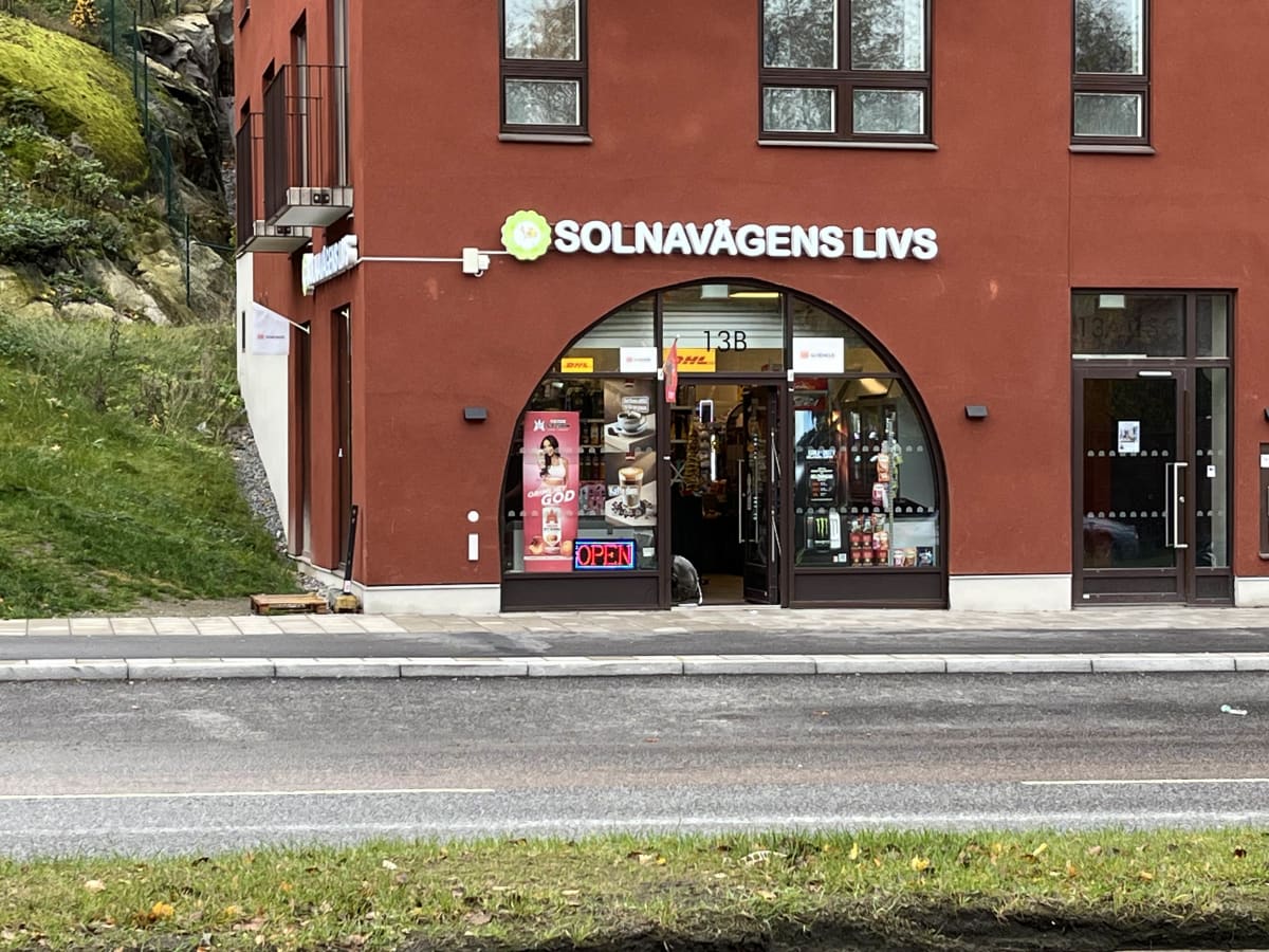 Bilden visar fasaden på en röd byggnad där en närbutik med namnet "SOLNAVÄGENS LIVS" är belägen. Butiken har en stor, välvd entrédel med glasdörrar som delvis står öppna. Inne i butiken syns hyllor med varor och olika reklamskyltar, inklusive en som annonserar "OPEN" med neonljus. Bredvid butiksentrén finns fönster och en dörr. Till vänster om butiken syns en grönskande sluttning med berg i bakgrunden. Framför byggnaden finns en trottoar och en asfalterad väg med en vit linje. Gräsband med några gula löv syns i förgrunden.