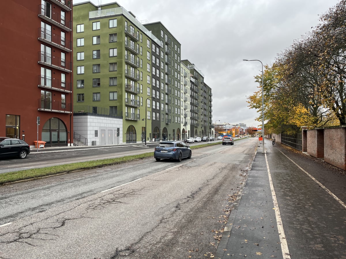 En ganska bred gata med flera hus i grön och röd färg. På vänster sida av bilden syns en röd byggnad med balkonger och fönster, och intill den står en grön byggnad. Längs gatan står det flera byggnader i varierande färger. Gatorna är asfalt med synli