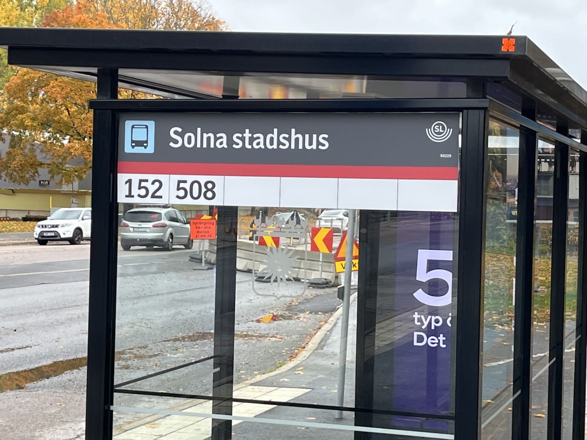 Bilden visar en busskur under dagtid, troligtvis under hösten då träden i bakgrunden har gula och orangea löv. Busskuren har ett mörkt tak och svarta stolpar. På busskurens skylt står det "Solna stadshus" med en bussikon, samt linjenummer "152" och "