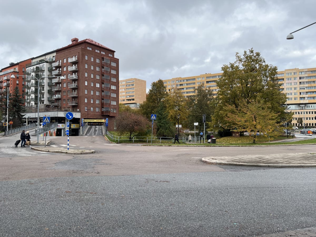 Bilden visar ett gatuområde med flera byggnader och träd. Till vänster syns bostadshus i rött och vitt med balkonger. Framför husen finns en nedfart till ett garage med skyltar som indikerar parkering och Solna Centrum. Två personer promenerar på tro