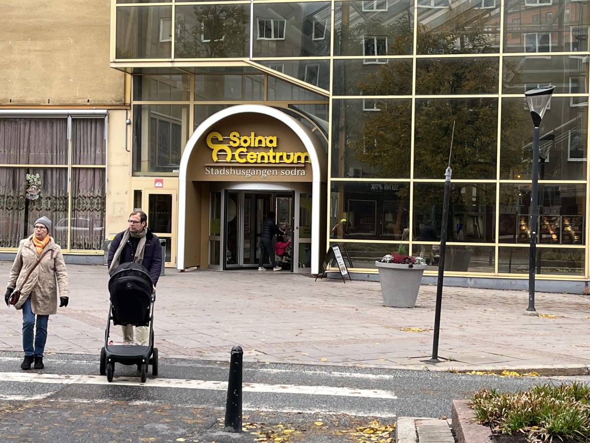Bilden visar entréområdet till "Solna Centrum" och "Stadshusgången södra" med flera personer i rörelse. Till vänster i bilden går en kvinna i beige kappa och orange halsduk, följt av en man som skjuter en barnvagn. Entrén till Solna Centrum består av