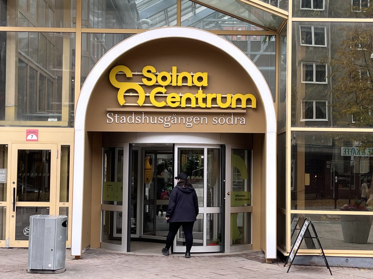 Bilden visar entréområdet till Solna Centrum, Stadshusgången södra. En stor gul skylt med texten "Solna Centrum" och "Stadshusgången södra" är placerad ovanför en automatisk karuselldörr och en intilliggande manuell dörr av glas och metall. En person sedd bakifrån, klädd i mörka kläder och keps, är på väg att gå in genom karuselldörren. Till vänster om entrén finns en grå metallkorgmarkerad med ett nummer '7' inuti en röd kvadrat. Ytterligare en dörr och större fönsterpartier syns på vänster sida. Till höger om entrén finns stora glaspartier som reflekterar omgivningen, inklusive en byggnad med fönster och träd med gulaktiga löv. En svart trottoarpratare står på marken framför högra glaspartiet. Golvet vid entrén är belagt med gatsten.