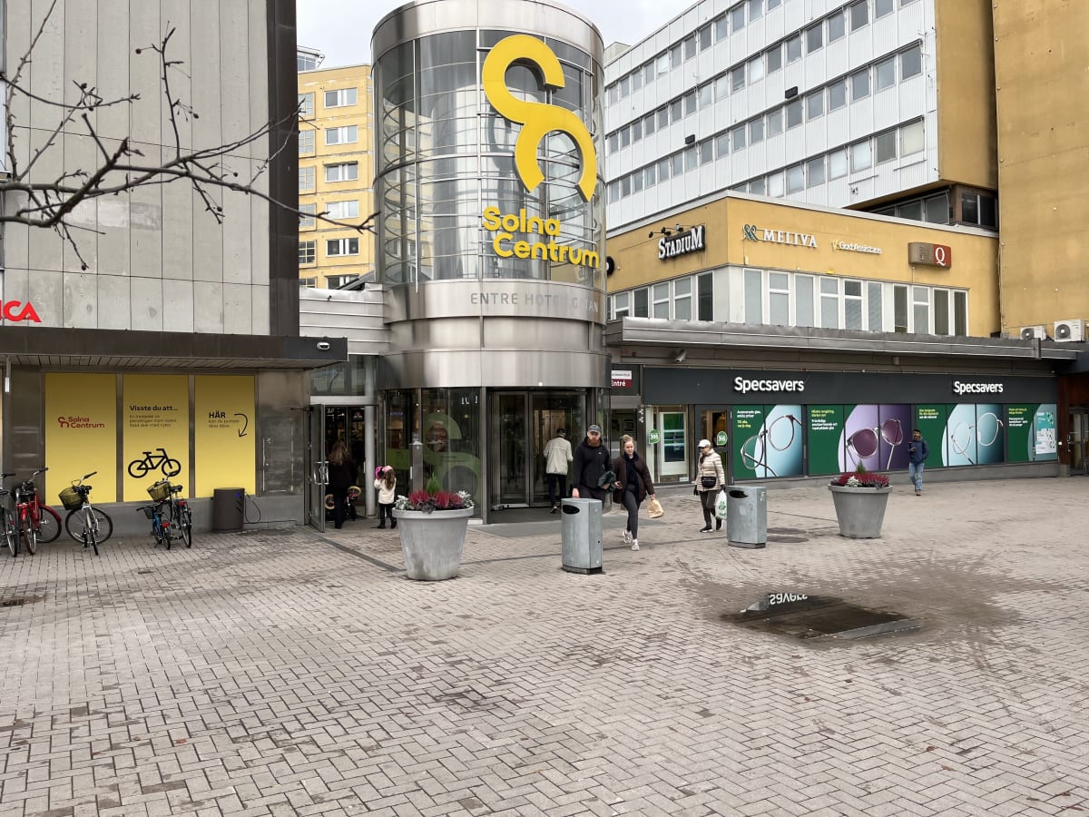 En bild tagen utomhus under dagtid, föreställande entrén till ett köpcentrum som heter 'Solna Centrum'. Entrén har en stor, rund glaskonstruktion med en gul Solna Centrum-logotyp. Människor går in och ut ur byggnaden. På vänster sida av entrén finns