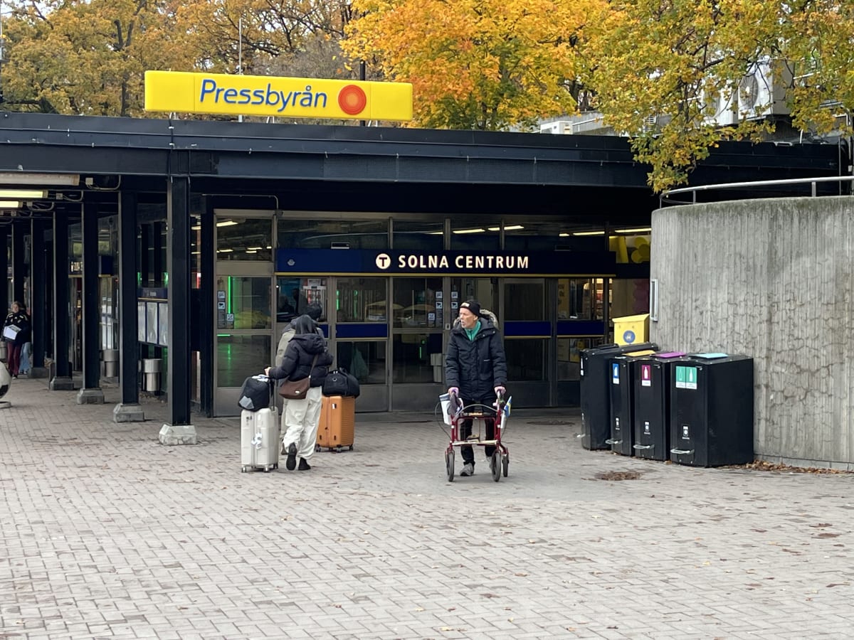 Bilden föreställer en utomhusscen framför vad som ser ut att vara ingången till en tunnelbanestation eller liknande offentlig byggnad. Ovanför ingången är en gul skylt med texten "Pressbyrån" och en röd symbol, samt en skylt med texten "SOLNA CENTRUM