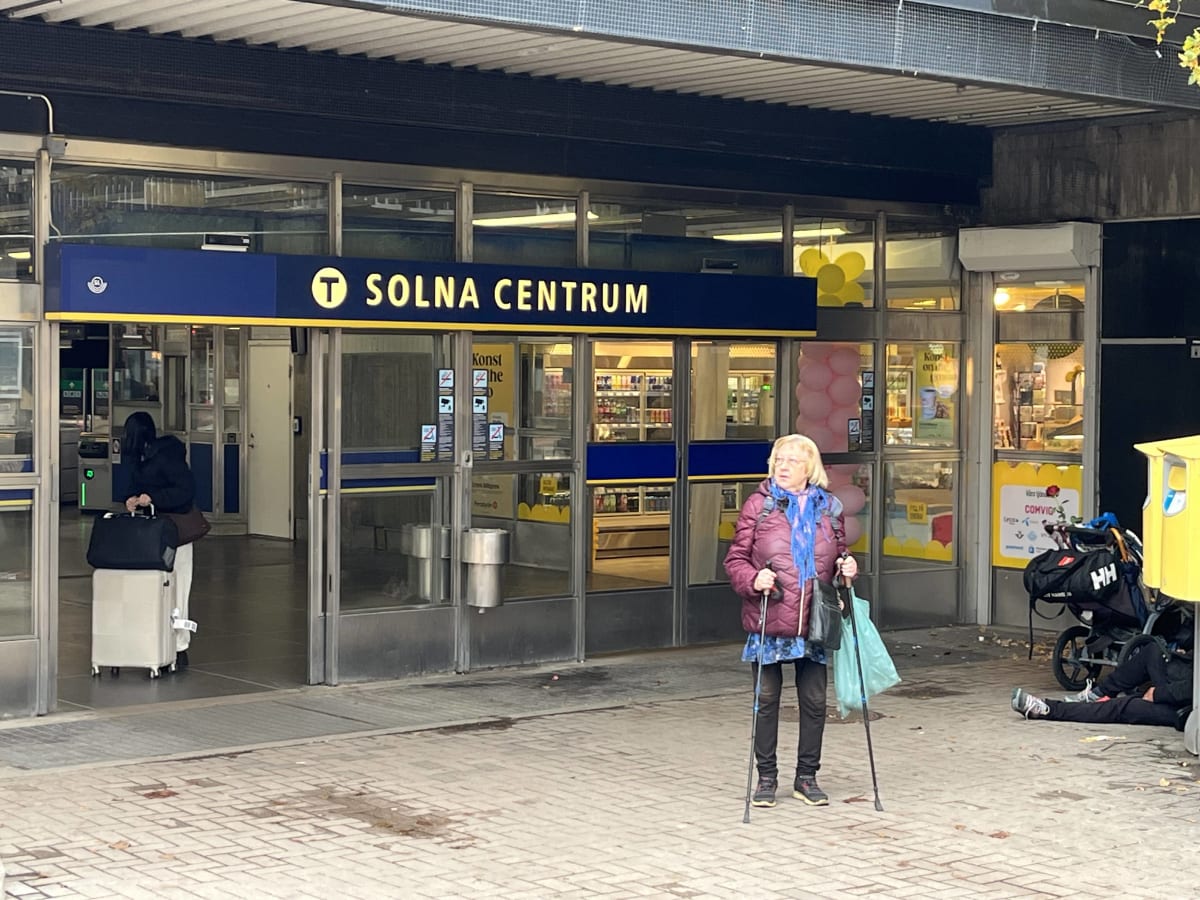 Bilden visar entréområdet till Solna Centrum, troligen en tunnelbanestation eller ett köpcentrum. Längst upp i mitten finns en stor blå skylt med texten "T SOLNA CENTRUM" i gult. Entrén består av dörrar i glas och metall, med flera affischer på dörra