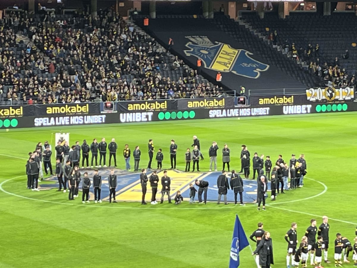 Bilden visar en sportarena, troligtvis en fotbollsarena, fylld med åskådare på läktaren i bakgrunden. En stor grupp människor, både vuxna och barn, står på gräsplanen i förgrunden, formerandes en cirkel eller halv-cirkel. Många av personerna på plane