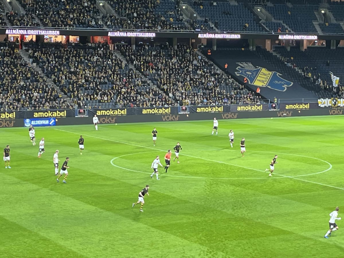En bild tagen från en läktare under en fotbollsmatch. På den gröna fotbollsplanen syns flera spelare i svarta och vita matchställ, samt en domare i orange tröja. Längs långsidan av planen finns reklamskyltar med texten "amokabel" och "voky", och ovan