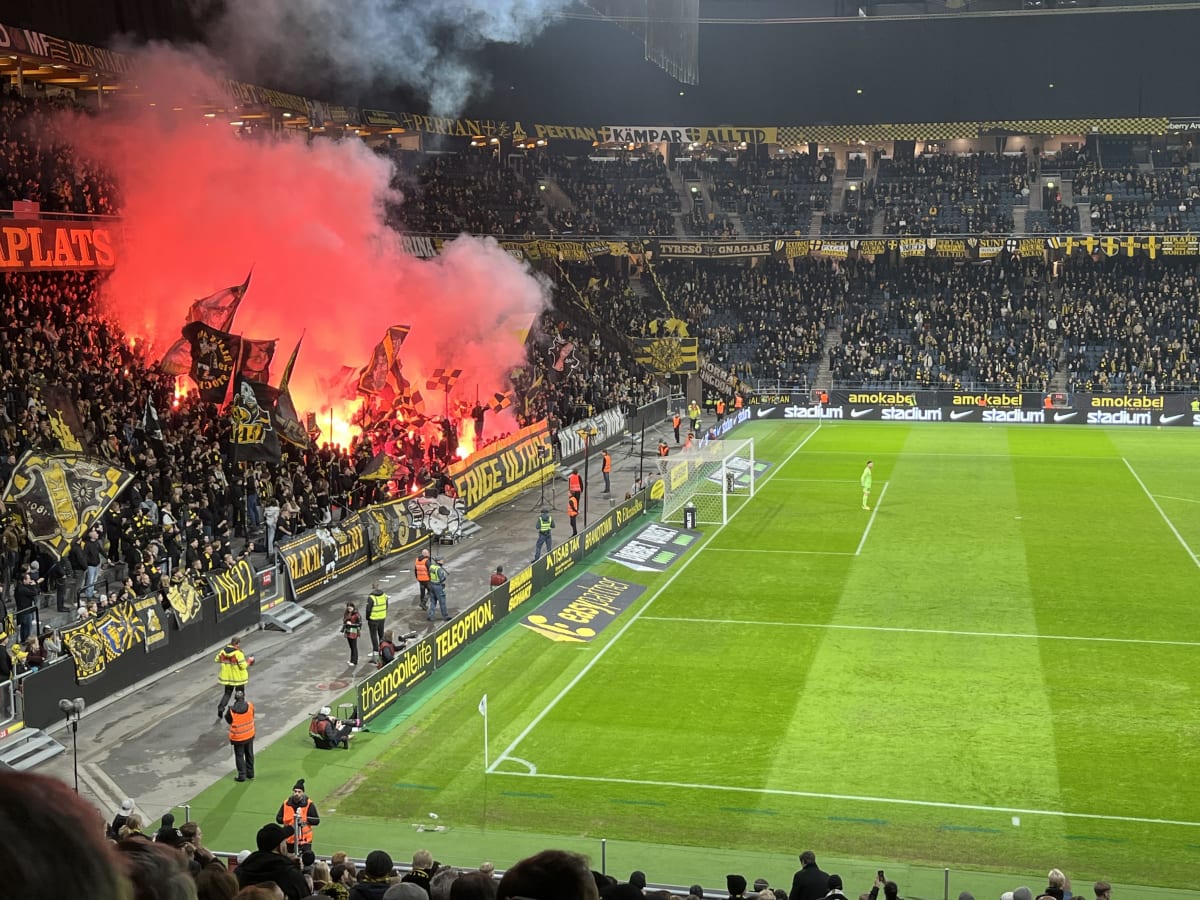 En vidvinkelbild tagen på en fotbollsarena under kvällstid, upplyst av artificiellt ljus. I den övre vänstra delen av bilden syns en sektion av publiken som håller upp flaggor och bengaler som avger röd rök, vilket skapar en stor röd rökpelare. Publi