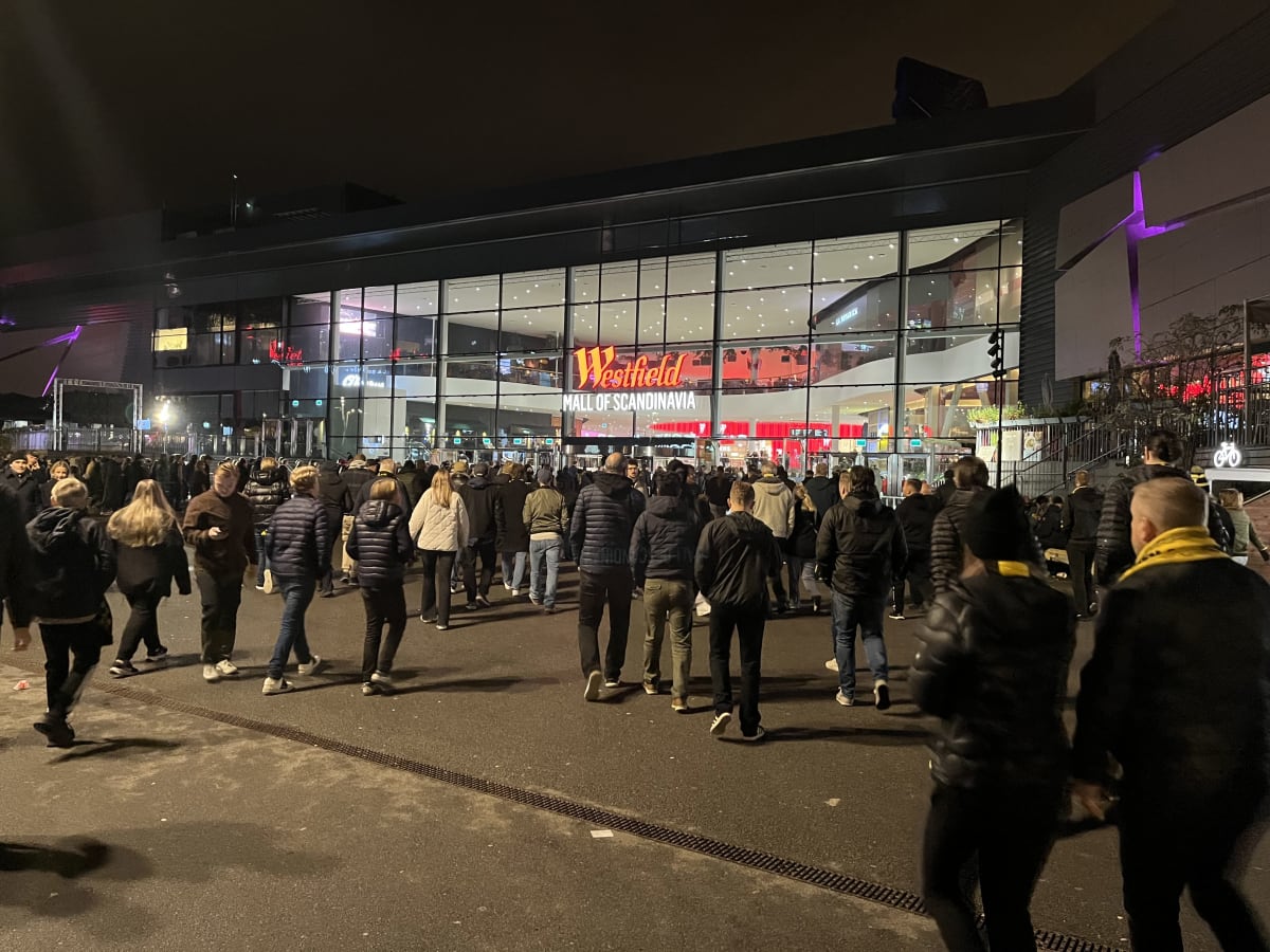 Bilden visar en folkmassa som står eller går framför entrén till ett stort köpcentrum som heter 'Westfield Mall of Scandinavia'. Det är natt och byggnaden är upplyst med fasadbelysning, inklusive en lila-aktig ljusdetalj på byggnadens högra sida. Fle