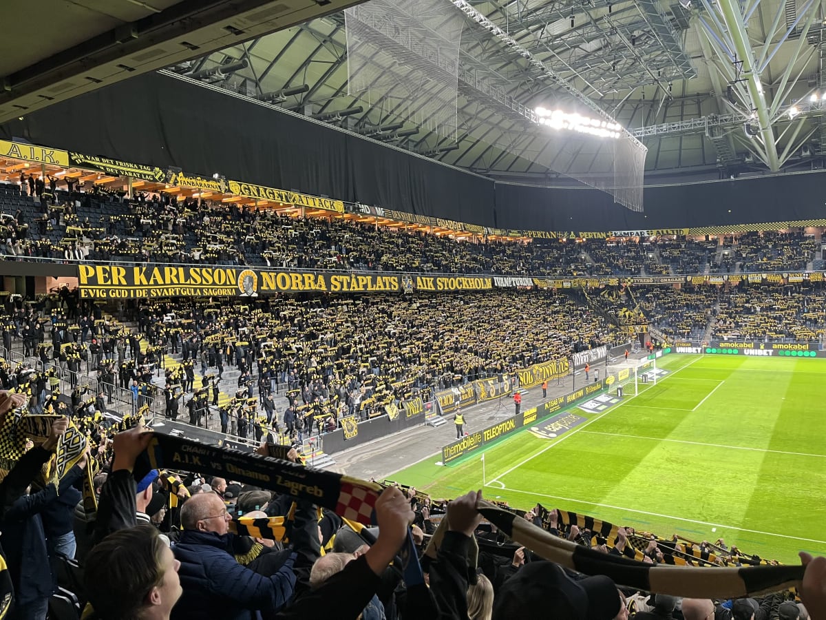 Bilden visar insidan av en stor inomhusarena eller fotbollsstadion fylld med publik. Större delen av åskådarna håller upp gula och svarta halsdukar eller tygstycken, vilket skapar ett enhetligt färgtäcke över läktarna. Flera banderoller syns på läkta