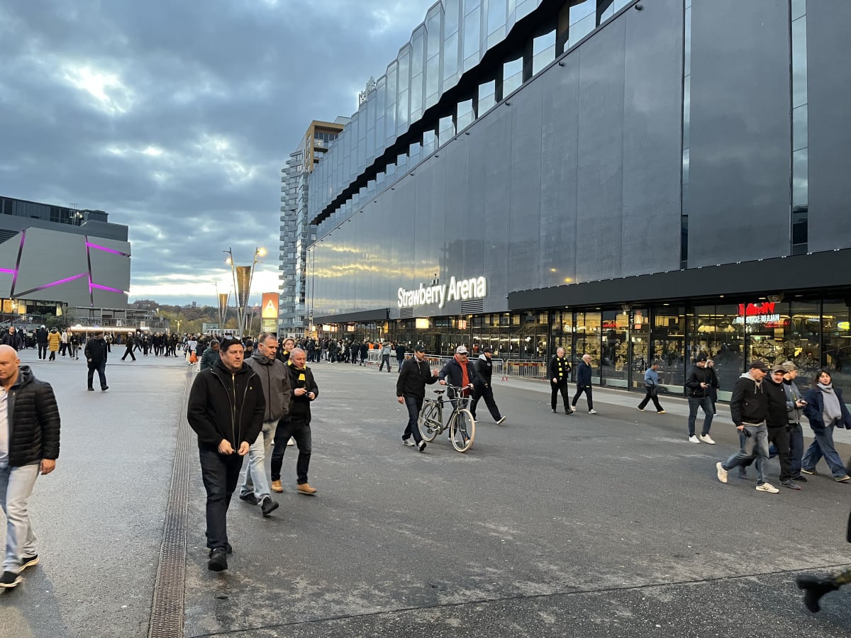En bild visar en folkmassa som rör sig på en asfalterad yta framför en stor byggnad. Byggnaden har namnet "Strawberry Arena" belyst på fasaden. Väggarna på byggnaden är gråa och har stora fönsterpartier som reflekterar himlen. En del av byggnaden har