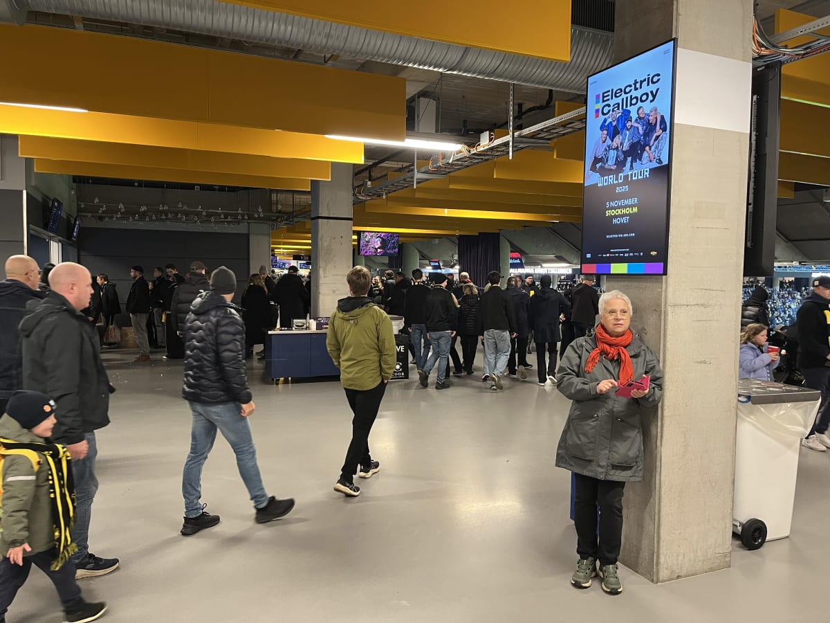 Bilden visar ett inomhusmiljö med en grupp människor som rör sig i en foajé eller korridor. Många av personerna är klädda i mörka ytterkläder. Till höger i bilden står en kvinna med grått hår och orange halsduk lutad mot en pelare. Hon håller i en ro