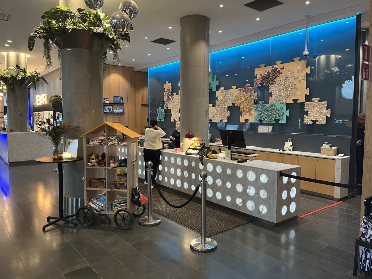 Bilden visar interiören av en modern lobby eller reception med en stor marmordisk i förgrunden, som har belysning som skapar vita cirklar på ytan. Bakom disken står två anställda, en vänd med ryggen mot kameran och en annan med ansiktet mot kameran.