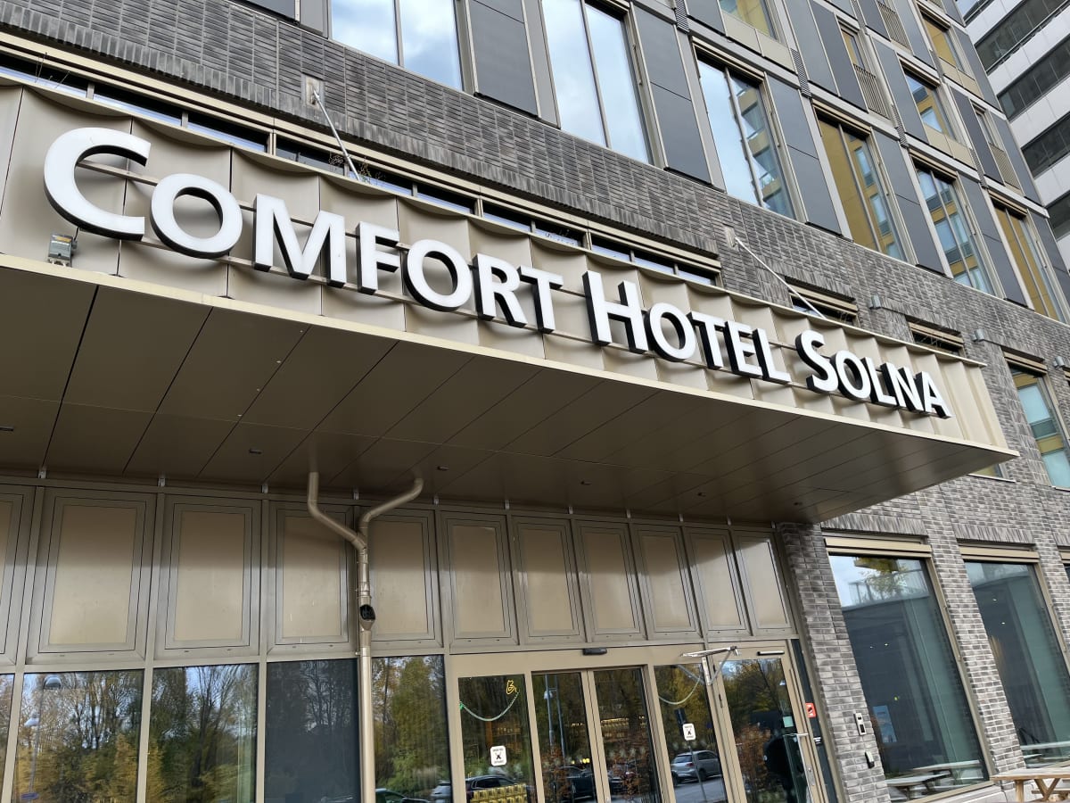 En bild tagen underifrån som visar entrén till "Comfort Hotel Solna". Ovanför en glasentré med dörrar finns en skylt med hotellets namn i stora, vita bokstäver på en ljusbrun gavel. Ovandelen av byggnaden består av fasadplattor i gråbruna nyanser och jämna rader av fönster med mörkt glas. Under skylten, direkt över entrédörrarna, finns ett avrinningsrör fastsatt på fasaden. I glasen reflekteras omgivningen, som inkluderar träd med glesa blad och en ljus himmel med moln.
