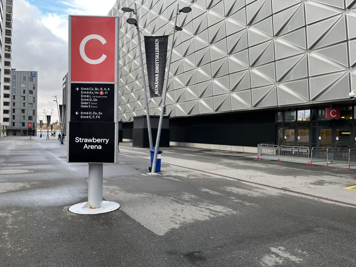 Bilden visar utsidan av en stor byggnad med en distinkt, mönstrad fasad bestående av ljusa, perforerade triangulära paneler. Framför byggnaden finns en asfalterad yta med en stor skylt i förgrunden. Skylten är röd och svart, visar en stor vit "C" överst och har texten "Strawberry Arena" längst ner med pilmarkeringar som pekar mot olika entréer. På skylten anges entréer som "Entré Cx, Bx, B, A, K", "Entré Kx, Hx", "Entré L Via Quality Hotel", "Entré G, Gx", "Gate 1 Entré Y", "Gate 2 Entré X", samt "Entré D, Dx, Ex" och "Entré E, F, FX". En svart flagga med texten "ALLMÄNNA IDROTTSKLUBBEN" är fäst vid en belysningsstolpe framför byggnaden. Längre bort syns en hög byggnad med många fönster och en logotyp för "Comfort Hotel". Vägytan framför byggnaden är delvis fuktig.