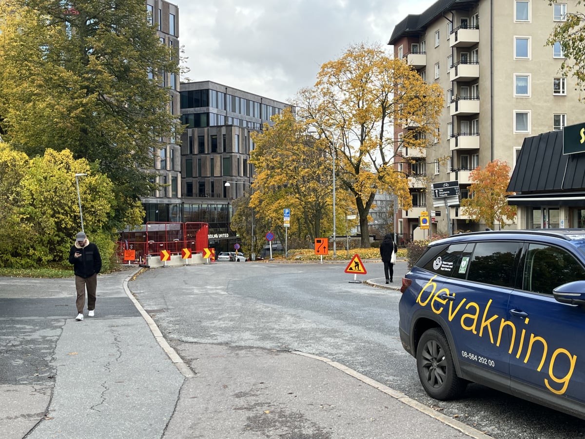 Bilden visar ett gatu- och byggnadsmiljö under dagtid. Till vänster i förgrunden går en person på en gångväg, klädd i mörka kläder och mössa, tittandes på sin telefon. I mitten syns en väg med avspärrningar och byggnadsställningar i rött, med gula pi