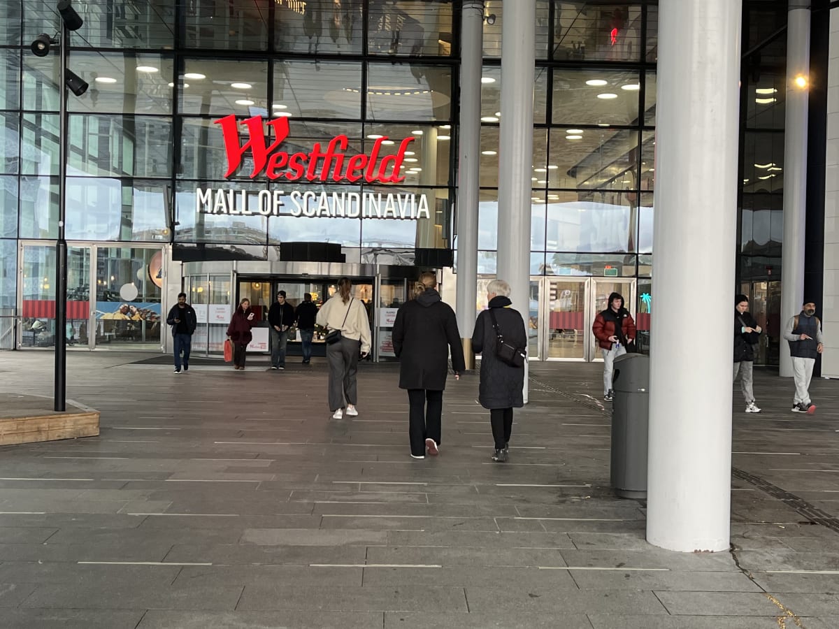 Bilden visar entréområdet till "Westfield Mall of Scandinavia". En stor glasfasad med texten "Westfield" i rött och "MALL OF SCANDINAVIA" i vitt är tydligt synlig. Flera personer, både män och kvinnor, befinner sig på den stenlagda ytan framför entré