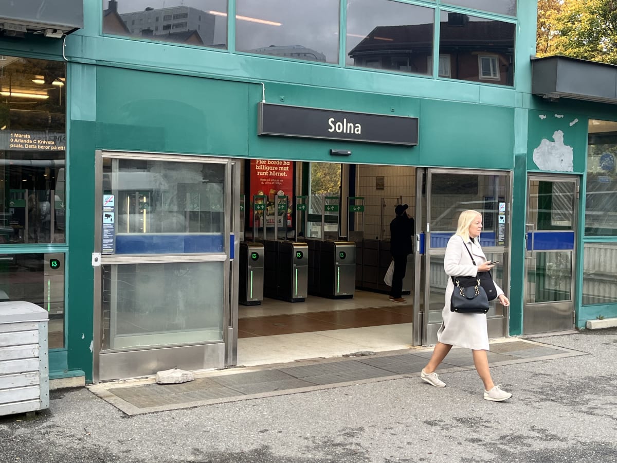 Bilden visar entrén till en tunnelbanestation eller pendelstation med namnet "Solna" skyltat ovanför ingången. Entrén är omgiven av en grön fasad med stora glaspartier som reflekterar byggnader och träd i bakgrunden. Ett par automatiska dörrar leder
