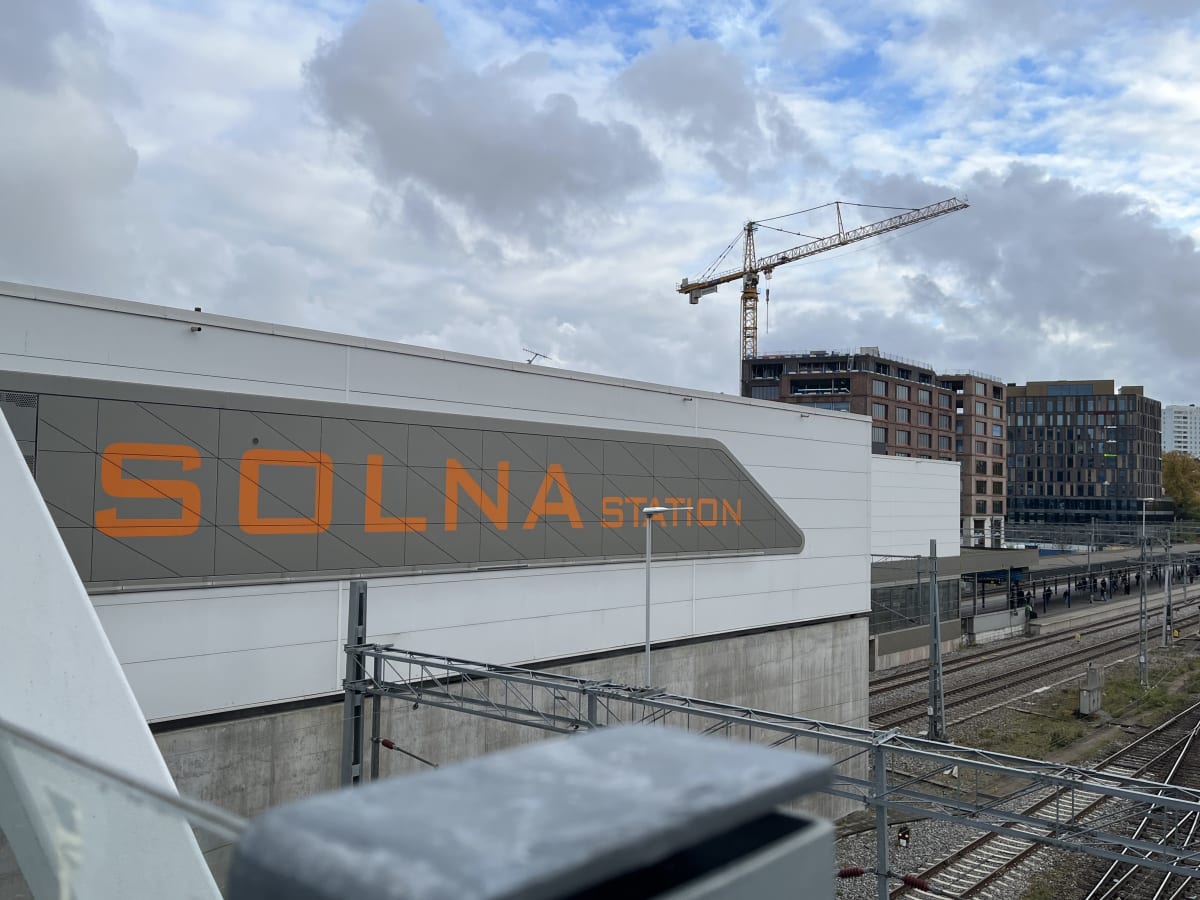 Bilden visar byggnaden 'Solna Station' med stor orange text på en grå fasad. En stor gul byggkran med texten 'SKANSKA' är synlig i bakgrunden, liksom flera andra byggnader, bland annat en med rödbrun tegelliknande fasad och en med mörkare fasad. Nedanför stationen finns flera tågspår med en plattform och ett fåtal människor. Elektricitetsledningar löper över tågspåren. Himmel med moln syns ovanför.