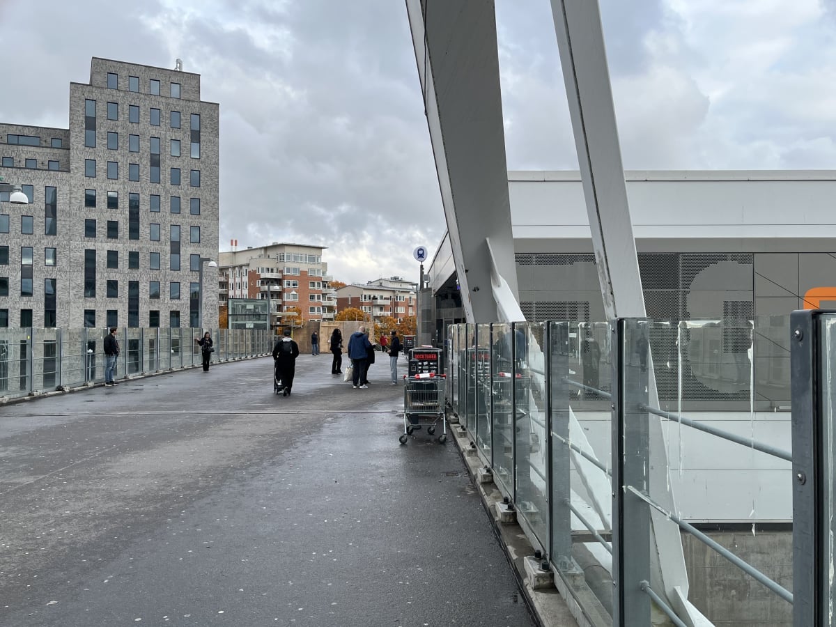 Bilden visar ett utomhusområde med flera byggnader under en mulen himmel. I förgrunden syns en bred gångväg där flera personer rör sig eller står. Till vänster på gångvägen står en man, och längre bort finns en kvinna som har lyft sin hand. I mitten