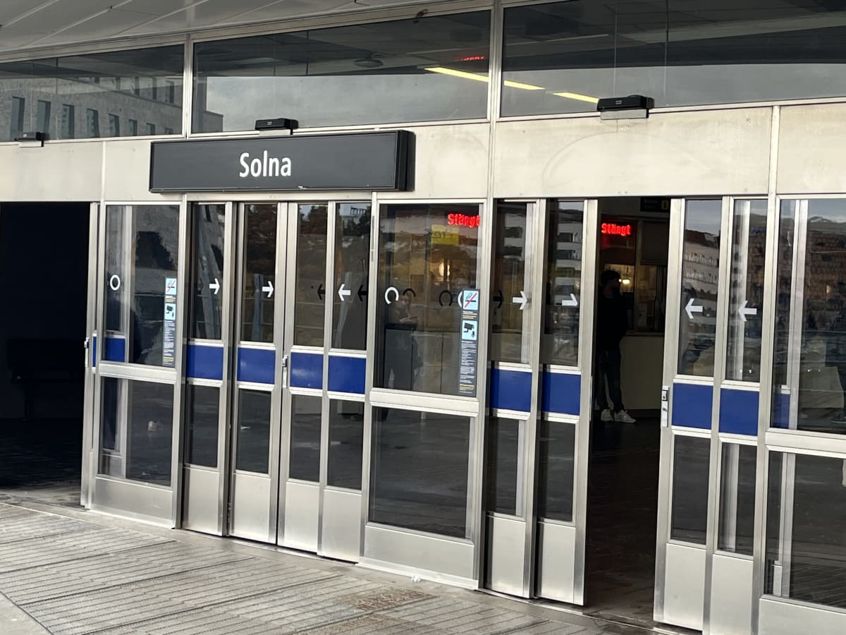 Bilden visar entrén till en byggnad, som framgår av skyltningen är 'Solna'. Entrén består av flera glasdörrar med silverfärgade ramar och blåa paneler i mitten. Över dörrarna finns en skylt med texten 'Solna' i vitt på svart bakgrund. En del av dörrarna har vita pilar som indikerar riktning. Reflektioner av en byggnad med fönster syns i glasdörrarnas övre delar. Innanför entrén syntes delvis en kassa eller informationsdisk, samt en person som står vid en öppen dörr. Röda digitala skyltar med texten 'Stängt' är synliga bakom glasdörrarna. Golvet framför entrén är ett räfflat grått golv. Ljuset indikerar att det är dagtid.