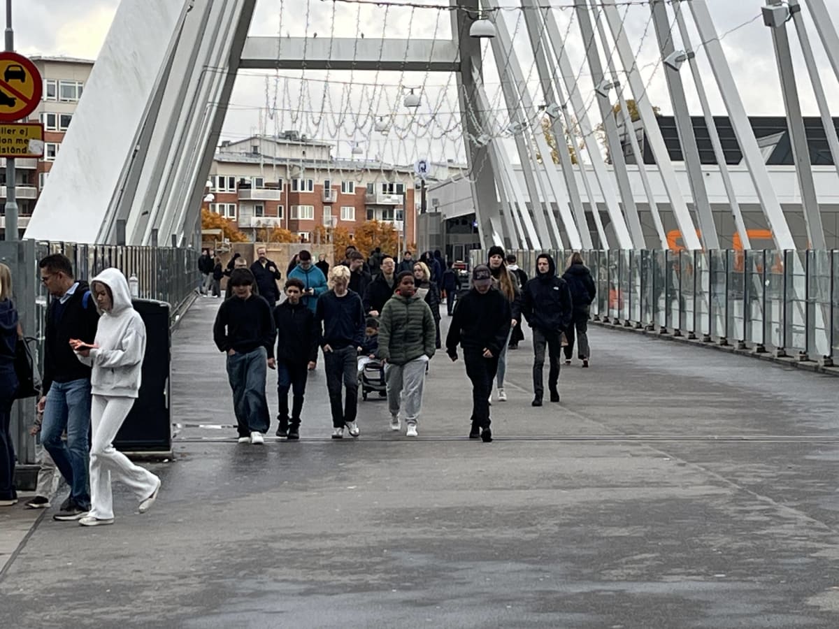 Bilden visar ett modernt gångstråk under en bro, möjligen en del av en större anläggning, där flera individer rör sig i olika riktningar. I förgrunden till vänster står en man och en kvinna, med en tredje person bakom dem. Kvinnan närmast kameran bär