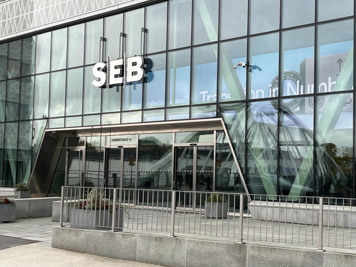 Bilden visar entréområdet till en modern glasbyggnad med texten "SEB" stort monterad på fasaden. Byggnaden har stora glaspartier som reflekterar omgivningen och en del av himlen. Vid entrén finns automatiska skjutdörrar. Framför byggnaden syns en grå stenläggning och flera avlånga planteringslådor med växter, samt ett metallräcke. På en av dörrarna syns en skylt med texten "STJÄRNFORGET 4". Inom glasfasaden kan man skymta text som "s bgroup m/NetZer a sition" och delar av vad som verkar vara en reklambild eller information med text och en del av ett kugghjulsliknande objekt. Två övervakningskameror är synliga på fasaden ovanför entrén.