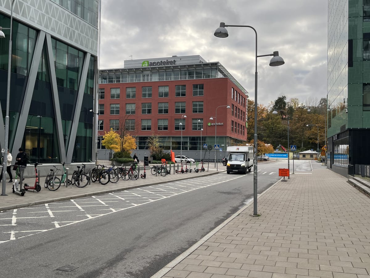 En gata med byggnader på båda sidor av bilden under ett molnigt himmel. Till vänster syns en modern kontorsbyggnad med glasfasad och geometriskt mönster. Till höger om vägen står en röd tegelbyggnad med många fönster och en grön skylt med texten 'apo