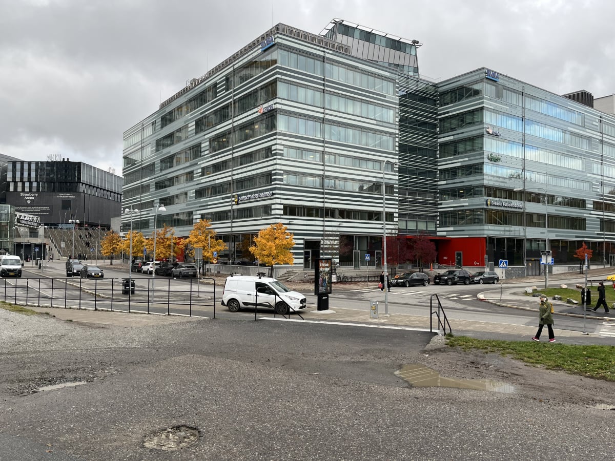 Bilden visar en stadsmiljö med flera moderna byggnader. I centrum står två stora, glasklädda byggnader med horisontella ränder i vitt och grått. På den vänstra av dessa byggnader syns en skylt med texten "Svenska Fotbollförbundet". Framför byggnadern