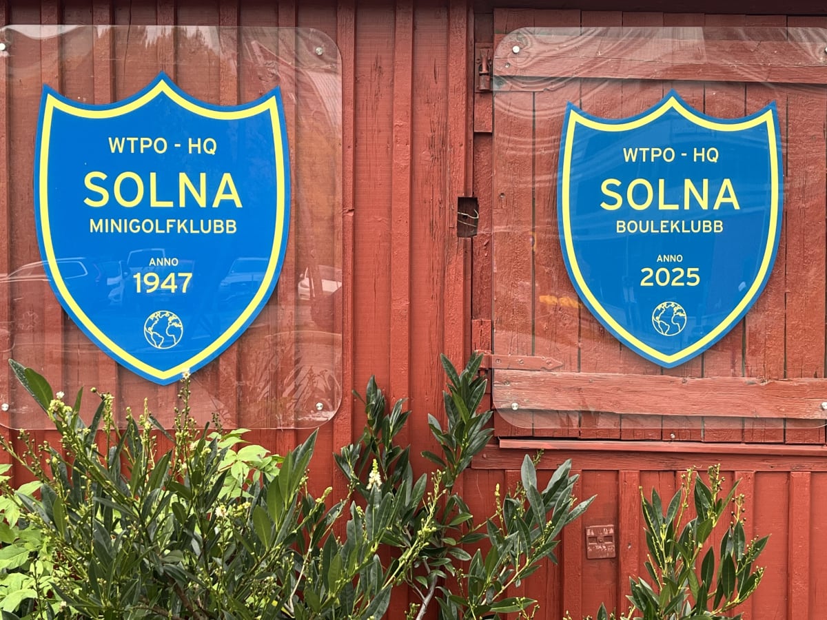 Bilden visar två blåa sköldformade skyltar med gul kant som är uppsatta på en röd träpanelvägg. Den vänstra skylten har texten "WTPO - HQ SOLNA MINIGOLFKUBB ANNO 1947" och en liten jordglob nedtill. Den högra skylten har texten "WTPO - HQ SOLNA BOULEKLUBB ANNO 2025" och en likadan jordglob. Skyltarna är monterade under transparenta plastark. Nedre delen av bilden domineras av gröna buskar med blad och små vita blommor eller knoppar. Vissa reflektioner av bilar kan anas på skyltarnas blanka ytor.