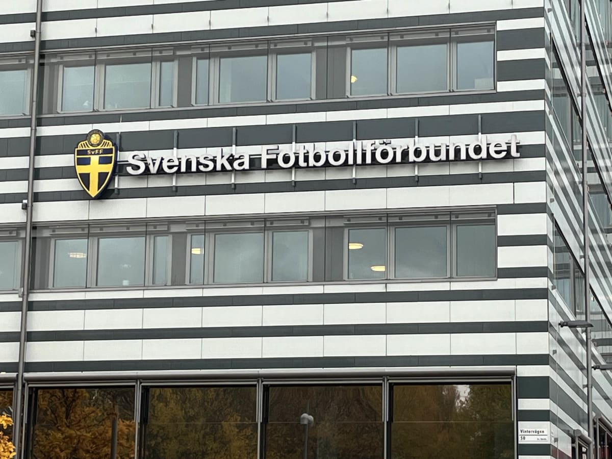 En bild tagen i en lätt uppåtvinklad position visar en modern byggnad med fasader av horisontella vita och grå-svarta ränder. På byggnadens framsida sitter skylten för 'Svenska Fotbollförbundet' med vita bokstäver och deras logotyp, ett gult och blått vapen med texten 'SVFF' och en fotboll överst. Skylten är placerad på den grå-svarta randen under en rad fönster och ovanför en annan rad med fönster. Delar av fasaden på byggnadens högra sida är gjord av glas som reflekterar omgivningen. Under den nedre raden av fönster på byggnadens framsida syns reflektioner av gula och orangea lövträd. En gatunamnskylt med texten 'Vintervägen' och '50' syns också i bildens nedre högra hörn.