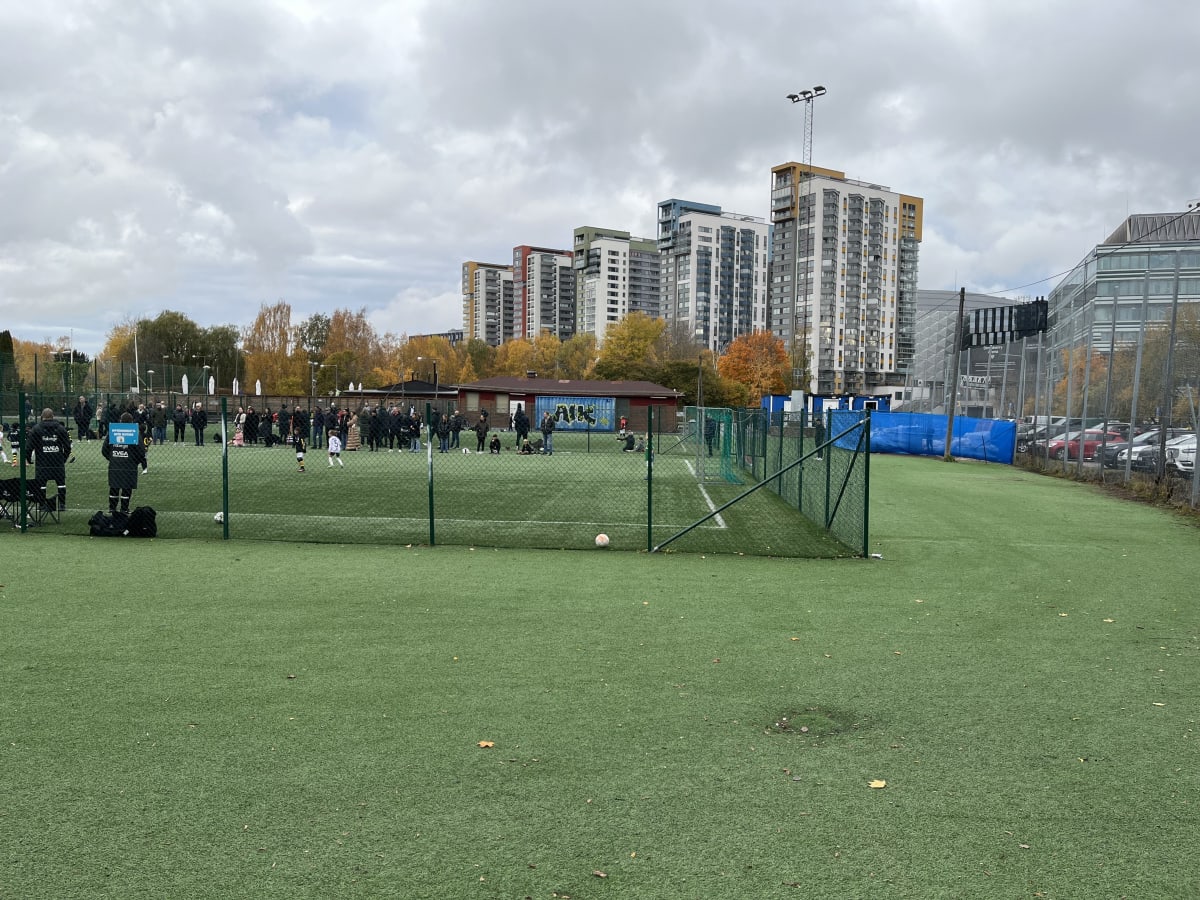 Bilden visar en utomhusmiljö med en grön konstgräsplan i förgrunden, omgiven av ett högt staket. Två vita fotbollar ligger på planen. Bortom staketet syns en grupp människor, av vilka några bär svarta kläder med logotyper som "Fabeg" och "SVEA". En b