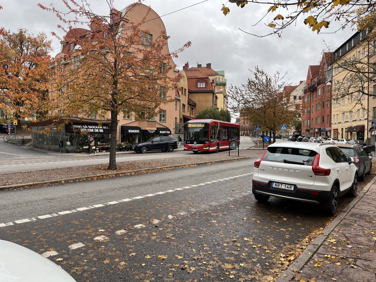 En ganska bred gatuvy på en höstdag, med ett flertal fordon och fotgängare. Asfalterade vägar är delvis täckta av gula och bruna löv. På höger sida av bilden syns bakre delen av en vit Volvo XC40 parkerad vid trottoarkanten. Längre fram på gatan, på