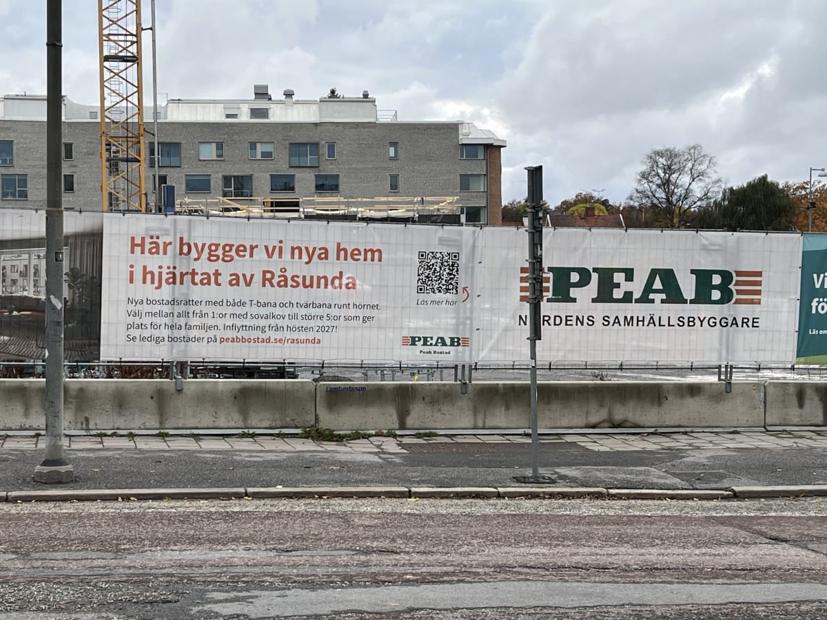 Bilden visar ett byggområde med en stor byggskylt i förgrunden som täcker den nedre delen av en byggnad. På skylten står det text om nybyggnation av bostäder i Råsunda, med information om storlekar, inflyttningsdatum och en QR-kod samt logotyp för Peab Bostad. Bakom skylten syns delvis en grå byggnad med fönster och en gul byggkran. I bakgrunden syns delar av träd med viss lövfällning och en gråmulen himmel. På marken framför skylten finns en asfalterad yta med trottoarkant och betongblock, samt en gatlykta eller liknande stolpe.