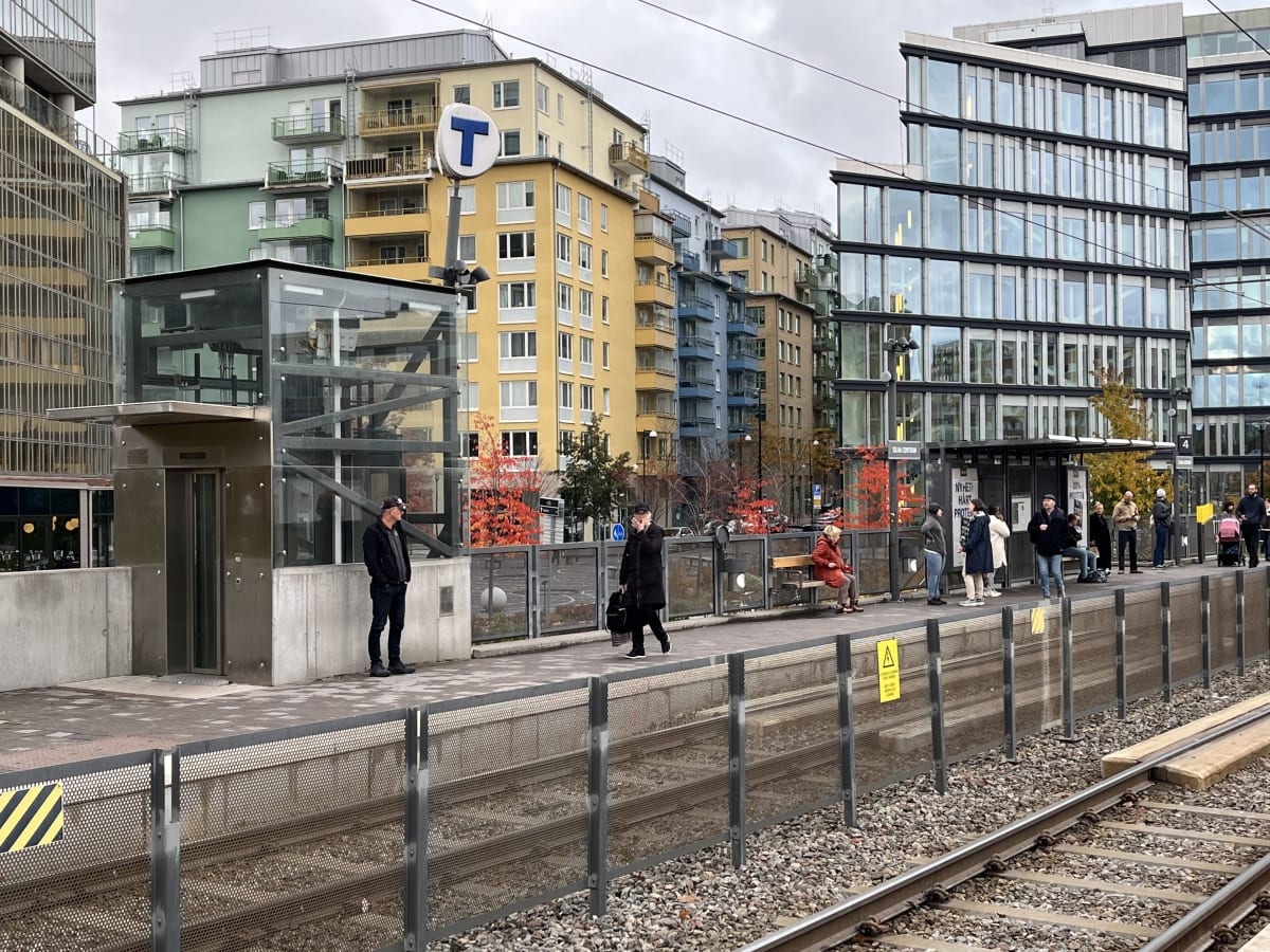 Bilden visar ett tågstopp eller en tunnelbanestation utomhus under dagtid med molnig himmel. I förgrunden syns tågräls med grå krossat grus mellan och bredvid dem en metallstaket som avgränsar perrongen. Staketet har varningstecken, bland annat ett g