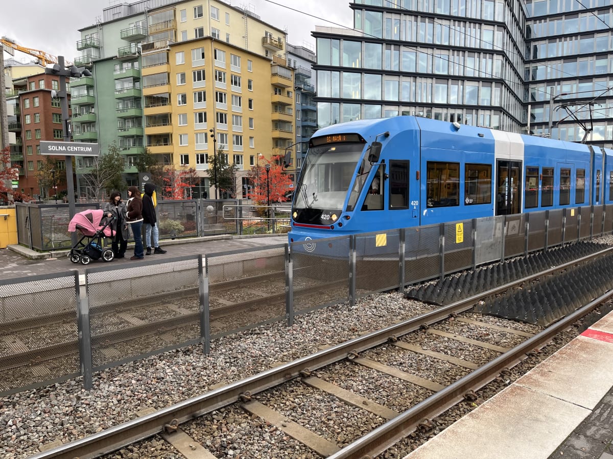 Bilden visar ett blått spårfordon vid en plattform. I förgrunden syns tågräls och en metallbur runt spåren. Tre personer, varav en med en rosa barnvagn, står på plattformen. En skylt med texten "SOLNA CENTRUM" är synlig. I bakgrunden finns flera bost