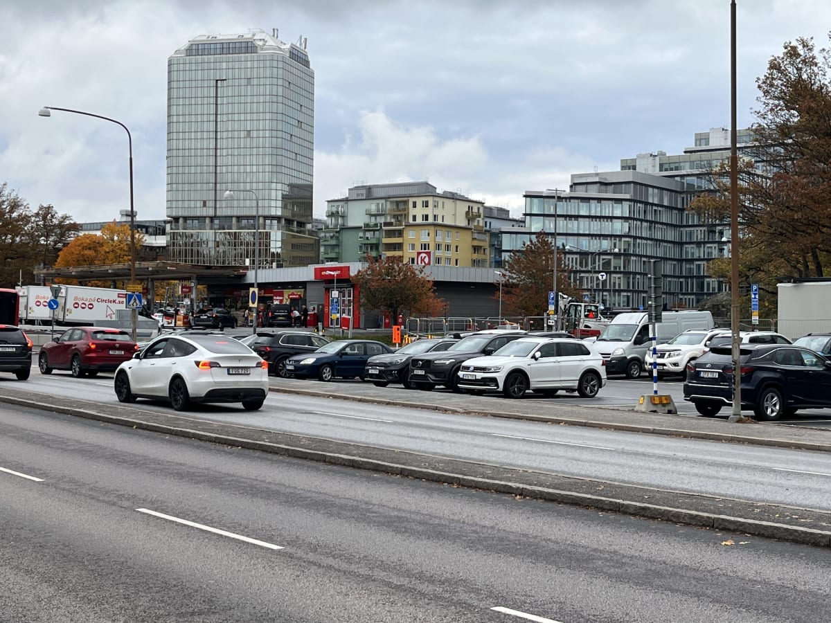 Bilden visar ett gatu- och stadslandskap med flera byggnader, fordon och en vägbana. I förgrunden syns asfalt med vita väglinjer och en trottoarkant. Ett flertal bilar är parkerade längs sidan av vägen, och ytterligare bilar rör sig på vägen, bland a