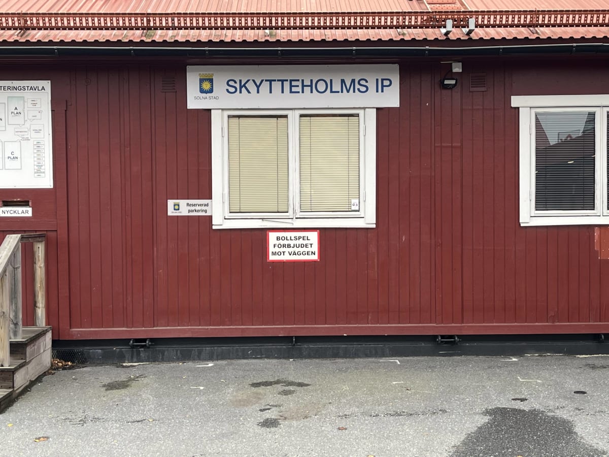 Bilden visar ett rött trähus med ett rödbrunt plåttak. På väggen sitter en vit skylt med texten "SKYTTEHOLMS IP" och texten "SOLNA STAD" tillsammans med Solna stads logotyp. Under denna skylt finns ett fönster med ljusa persienner. Till höger om fönstret finns en svart säkerhetskamera eller belysning monterad på väggen. Till vänster om fönstret sitter en mindre skylt som säger "Reserverad parkering". Under fönstret sitter en röd och vit skylt med texten "BOLLSPEL FÖRBJUDET MOT VÄGGEN". Till vänster i bilden syns en del av en trappa av trä. Markytan framför byggnaden är asfalt med några löv och mörka fläckar.