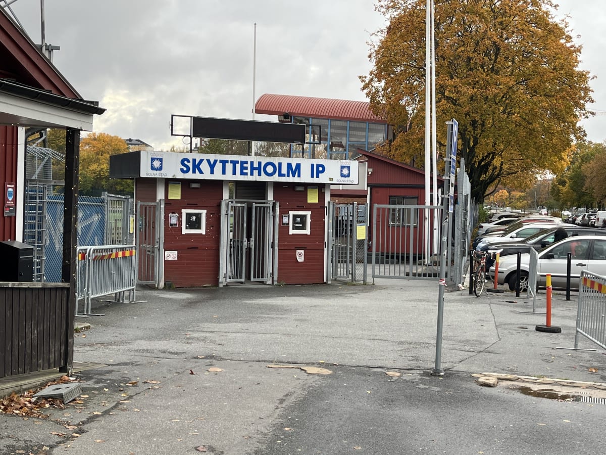 Bilden visar entréområdet till 'SKYTTEHOLM IP' en mulen dag. I mitten av bilden finns en röd byggnad med blå text som markerar platsen. Ingången består av ett antal grå metallgrindar och vändkors. Framför byggnaden finns en asfalterad yta med en del löv och skräp på marken. Till höger om entrébyggnaden finns en parkering med flera bilar uppradade. I bakgrunden syns träd med löv i höstfärger, samt delar av byggnader och den grå himlen. På skylten ovanför entrén syns även en logotyp för 'SOLNA STAD'. På entrébyggnaden sitter olika skyltar och en brevlåda är monterad på vänster byggnadsdel.
