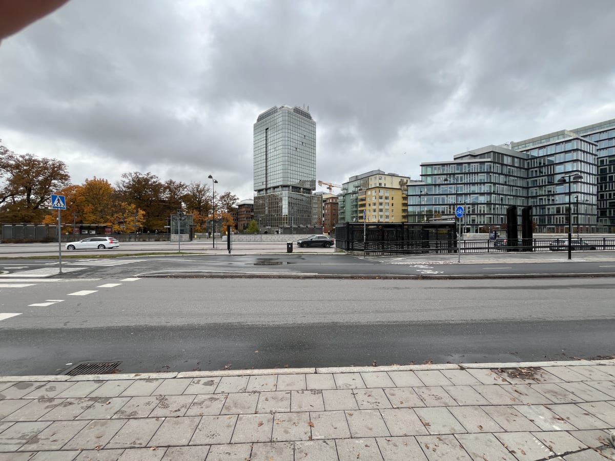 Bilden visar ett stadslandskap under en molnig dag. I förgrunden syns trottoar och en våt vägbana med vita markeringar, inklusive ett övergångsställe. På vänster sida av vägen syns ett vitt fordon, som ser ut att vara en kombibil, följt av höstlövsklädda träd i nyanser av orange och brunt. Ett flertal byggnader syns i bakgrunden, däribland en hög, modern kontorsbyggnad med glasfasad och flera flerbostadshus i varierande färger som gult och grått. Det finns även busshållplatser, skyltar (inklusive en för övergångsställe och en för cykelbana) och gatlyktor. En svart kombibil står parkerad vid trottoarkanten till höger i bilden. Mellan byggnaderna anas en gul byggkran.