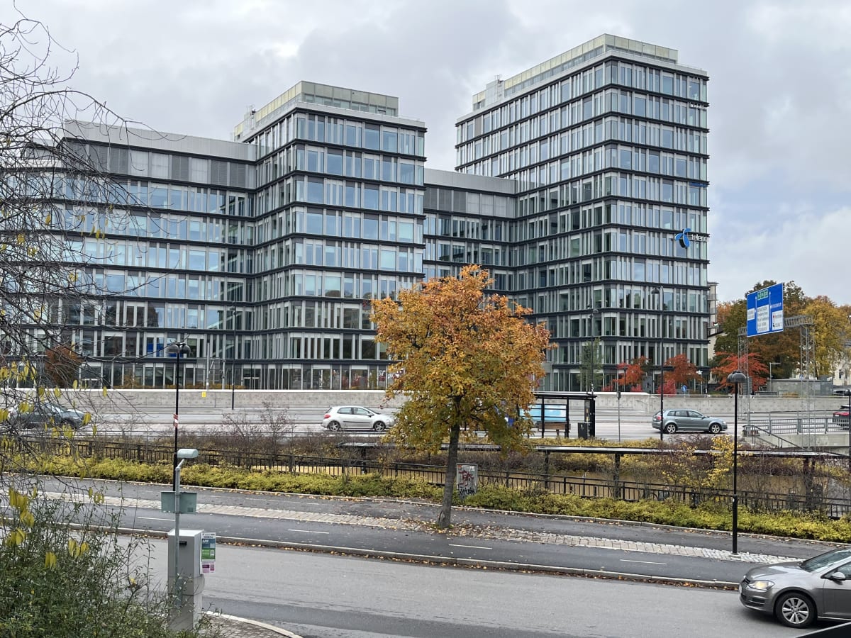 En bild som visar en modern kontorsbyggnad med många glasfönster. Framför byggnaden finns en väg med flera körfält där några bilar kör, samt en trädrad och en busshållplats. Träden har höstfärger i gula och orangea nyanser. Himlen är mulen och grå. I förgrunden syns ett flertal trädgrenar med spridda gula löv. Till vänster i bilden syns en parkeringsmätare eller liknande stolpe med en skylt som uppmanar till att använda en app. Mot den högra delen av bilden syns en blå vägskylt med vit text.