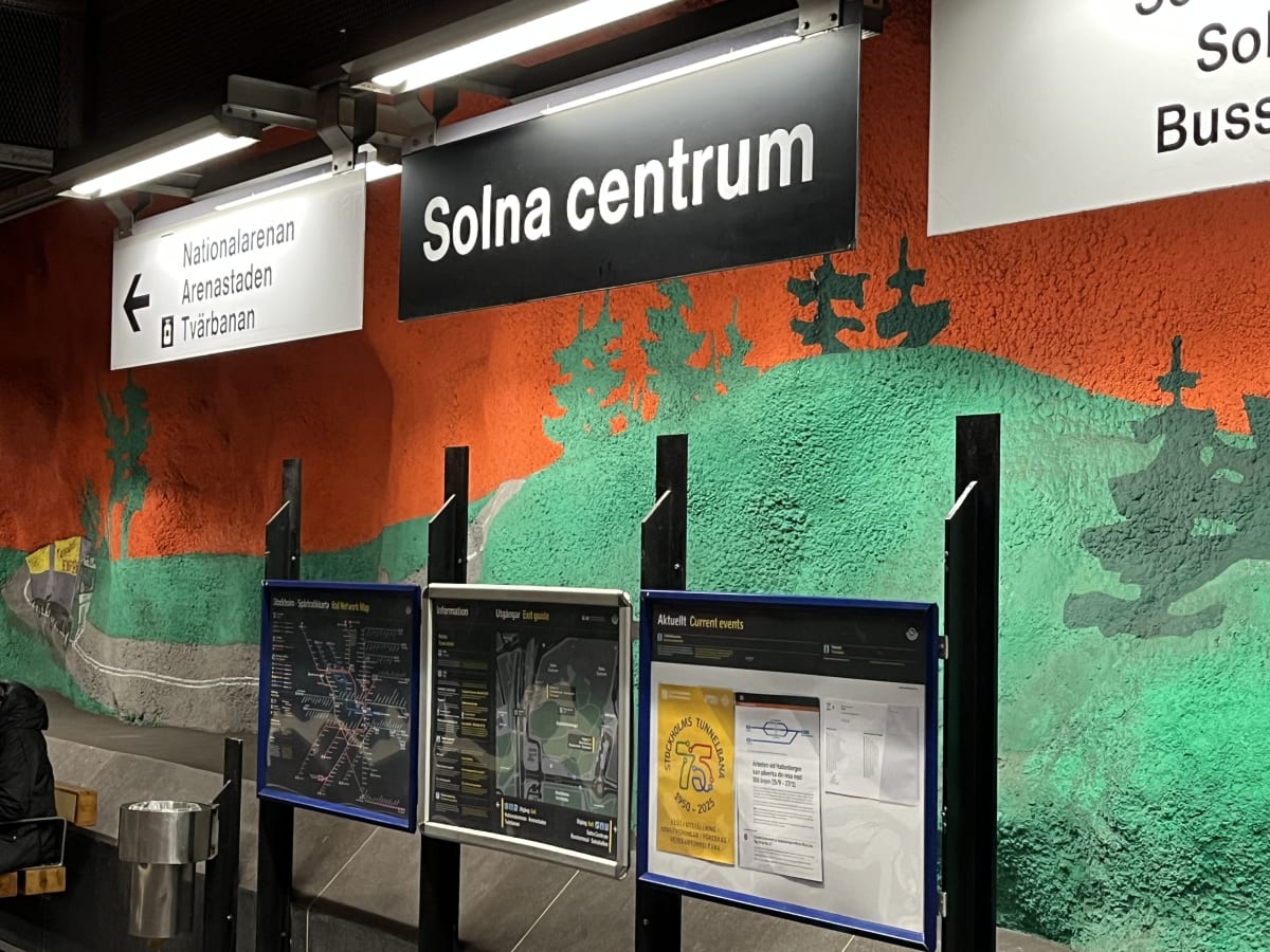 Bilden visar en del av en tunnelbanestation. I förgrunden syns ett par skyltar som pekar mot olika riktningar, bland annat "Nationalarenan", "Arenastaden" och "Tvärbanan". Ovanför finns en stor svart skylt med texten "Solna centrum" i vitt. Väggen bakom skyltarna är täckt av en detaljerad väggmålning i orange och grönt, som föreställer ett landskap med träd och vad som ser ut som en väg eller flod. Under skyltarna finns tre informationstavlor som visar en karta över Stockholms spårtrafiknät, en utgångsguide och en tavla märkt "Aktuellt Current events" med information och annonser, bland annat en affisch för "Stockholms Tunnelbana 75 år". Till vänster i bild syns också en sittbänk med en del av en persons ben och en papperskorg i metall.