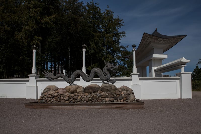 Bilden visar ett landskap med en konstgjord installation i förgrunden. I mitten av bilden finns en stor staty av en ormliknande drake, troligen i metall, som ringlar sig över ett fundament av stenar. Bakom statyn sträcker sig en vit mur prydd med kol
