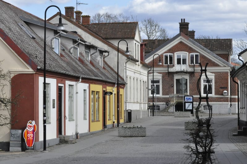 Bilden visar en gata med kullersten kantad av byggnader med olika fasadfärger, inklusive rött, vitt och gult. Det finns flera fönster och dörrar synliga på byggnaderna. På vänster sida av gatan står en röd figur som liknar en glassförsäljare, tillsam