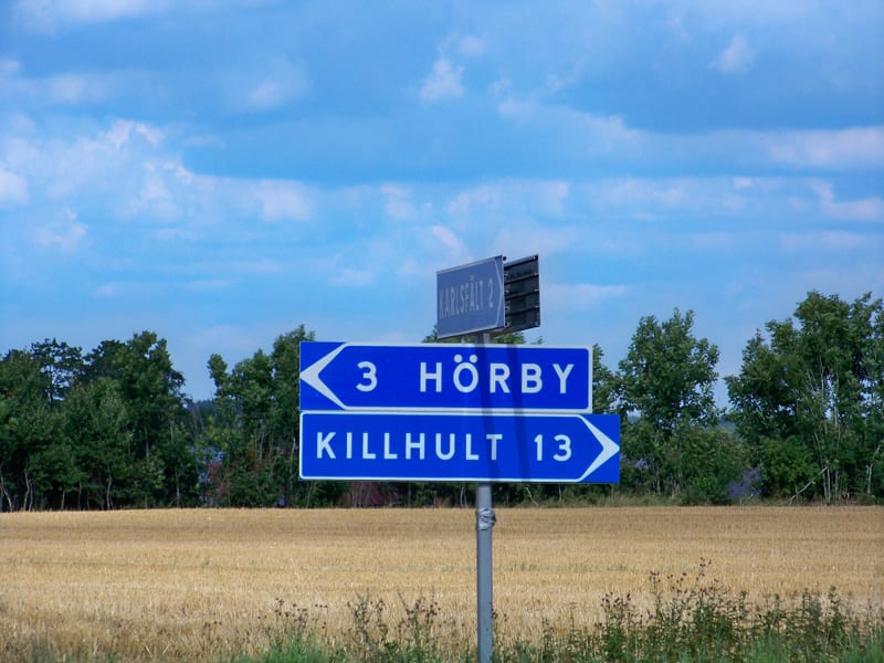 Bilden visar flera blå vägskyltar på en grå stolpe. De två större skyltarna visar riktning mot "HÖRBY 3" och "KILLHULT 13" med vita versaler och vänsterpekande pilar för Hörby och högerpekande pilar för Killhult. En mindre, mörkare skylt med texten "