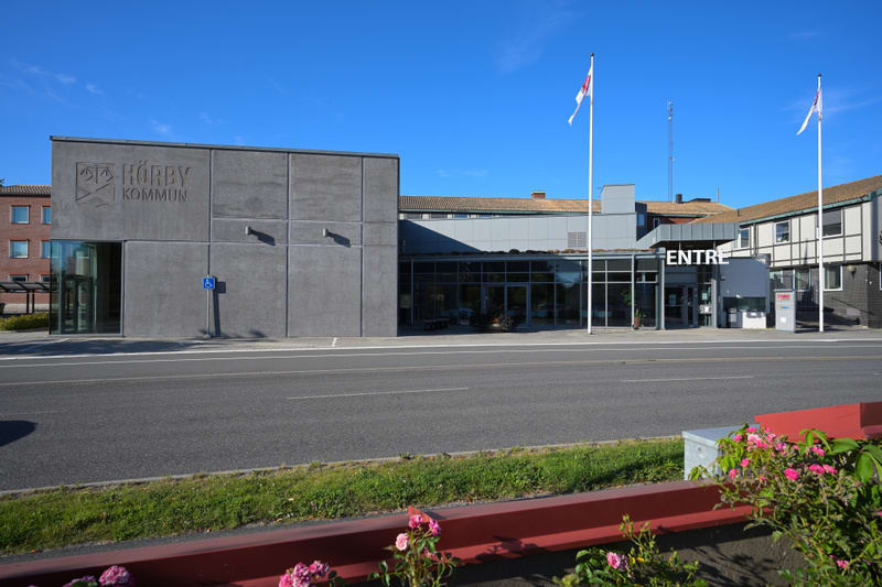 En fasadvy av en kommunkontorsbyggnad under dagtid. Byggnadens vänstra del är en grå betongkonstruktion med texten 'HÖRBY KOMMUN' ingraverad, samt en symbol och en skylt som indikerar tillgänglighet för rullstolar. En ingång med glasdörrar finns till