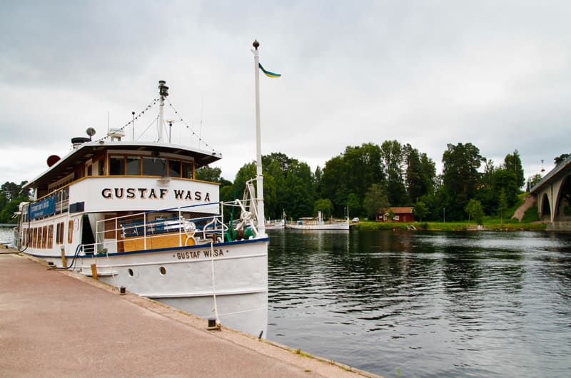Bilden visar ett vitt fartyg med namnet "GUSTAF WASA" vid en kaj på vänster sida. Fartyget har en blå och gul flagga på en flaggstång. Till höger om fartyget syns en vattenyta som leder bort i bilden. På motsatt sida av vattnet finns en grönskande st