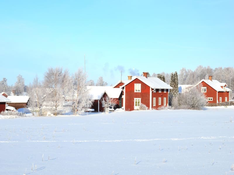 Bilden visar ett vinterlandskap med röda hus under en klarblå himmel. I förgrunden syns ett snötäckt fält. Bakom fältet står flera traditionella röda trähus med vita knutar och snötäckta tak. Från en skorsten på ett av husen stiger rök upp mot himlen